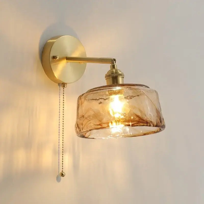 A warm amber glass copper led wall lamp with textured design and brass finish for bedroom use with pull chain