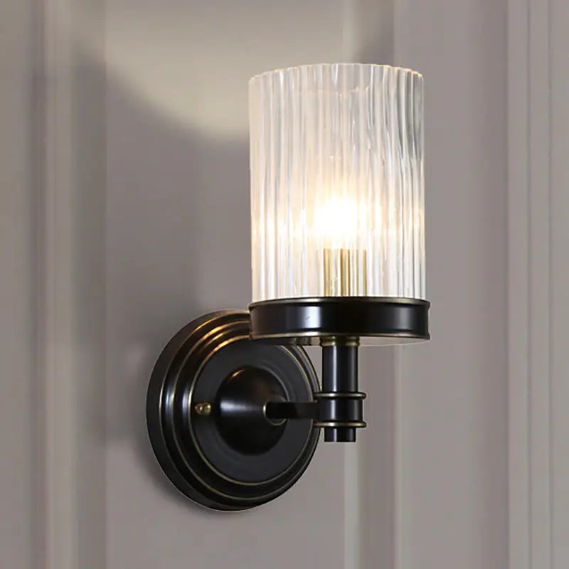 Vintage brass and black wall lamp with clear ribbed glass cylinder shade emitting warm light for bedroom use