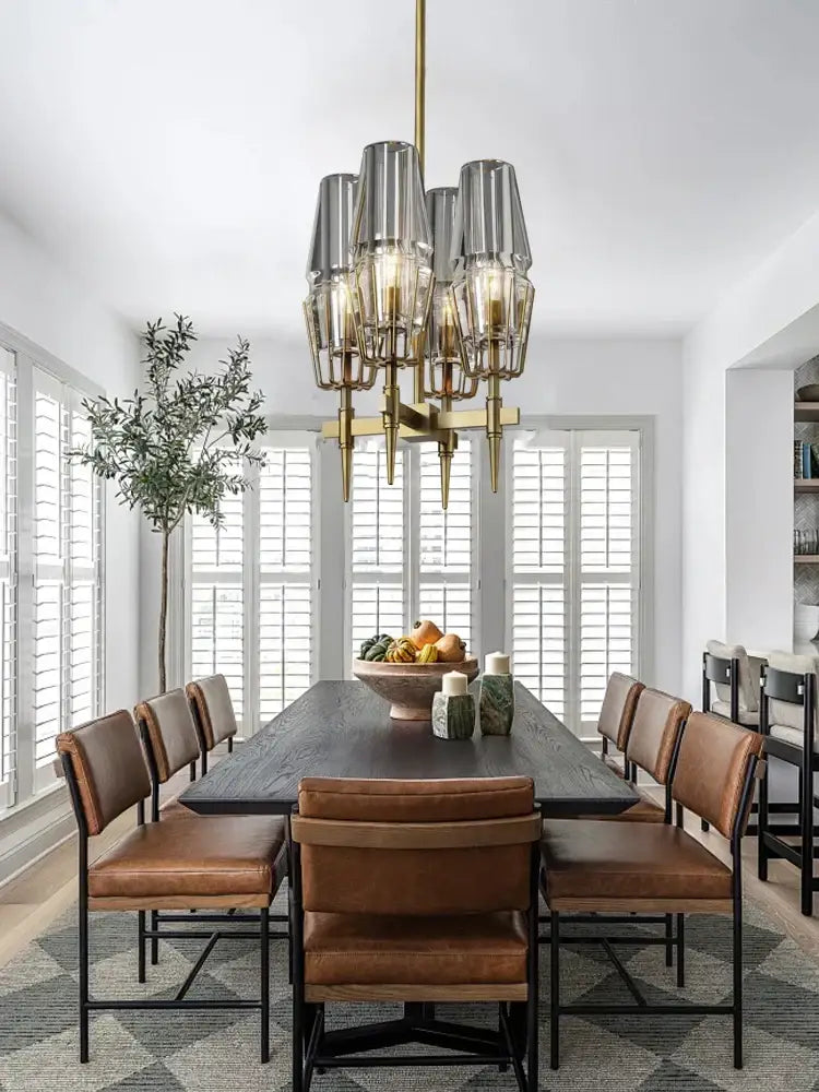 A modern decorbites brass crystal chandelier with clear glass shades hangs above a dining table with smoked glass accents