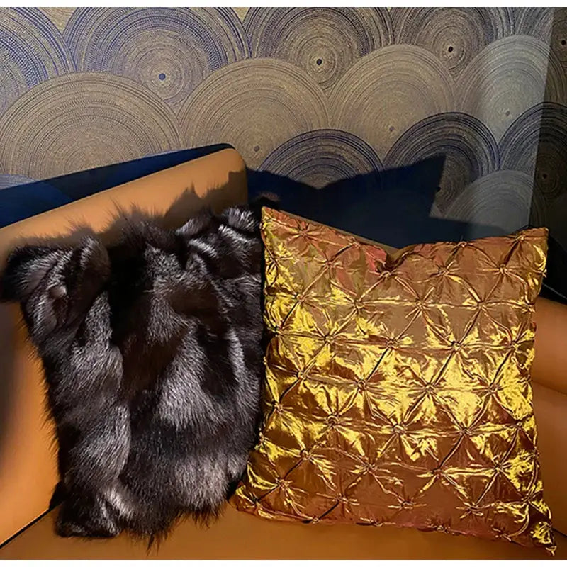 A luxurious gold-toned satin cushion cover with quilted pattern beside a black decorbites fox fur cushion on orange couch