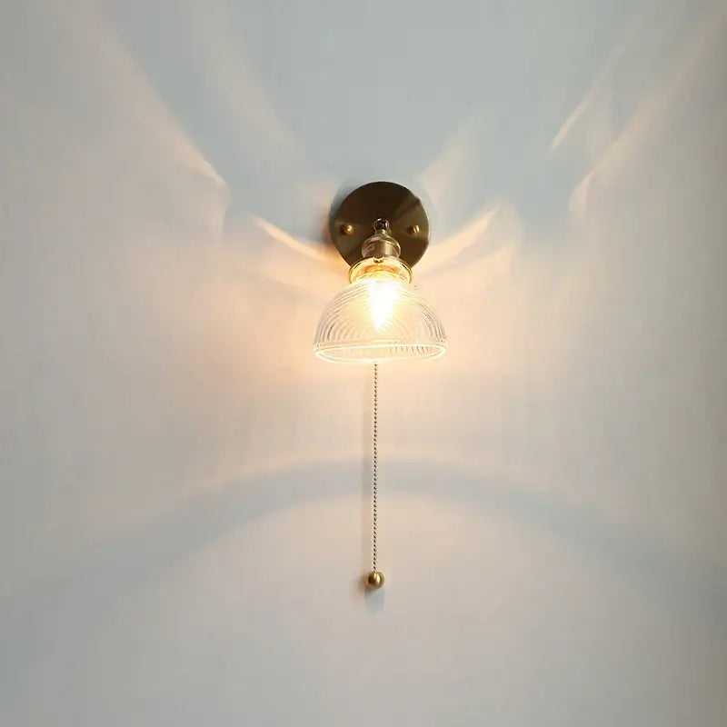 A warm-toned brass wall sconce with clear ribbed glass shade and ornate pull chain for vintage green glass led lighting