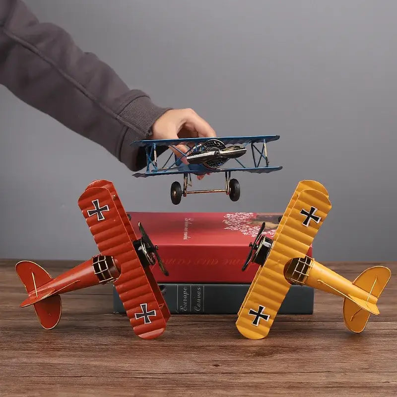 A hand holds a blue metal airplane figurine with black wheels above vintage red and yellow decorbites iron airplane models on books