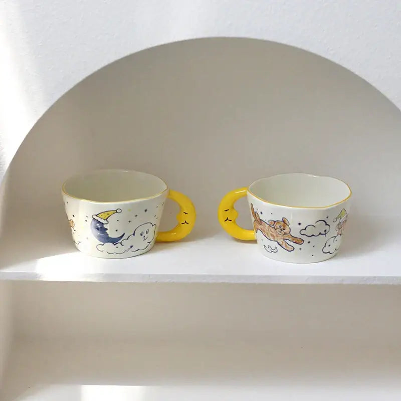 Two whimsical off-white bear moon ceramic mugs with yellow handles and hand-painted moon and cat illustrations