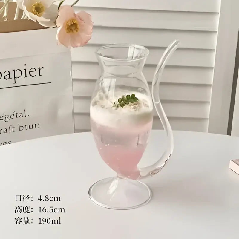 A clear glass cocktail glass cup with curved handle filled with pink vampire cocktail glass garnished with green herbs