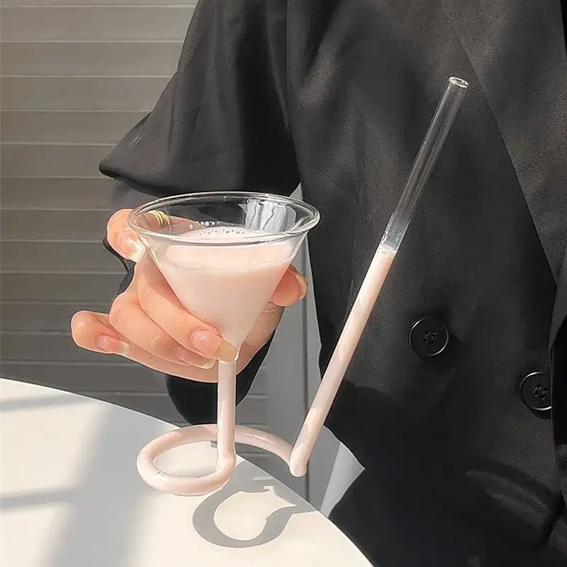 A clear glass vampire cocktail glass cup with a pink curved stem and handle holding a light pink beverage