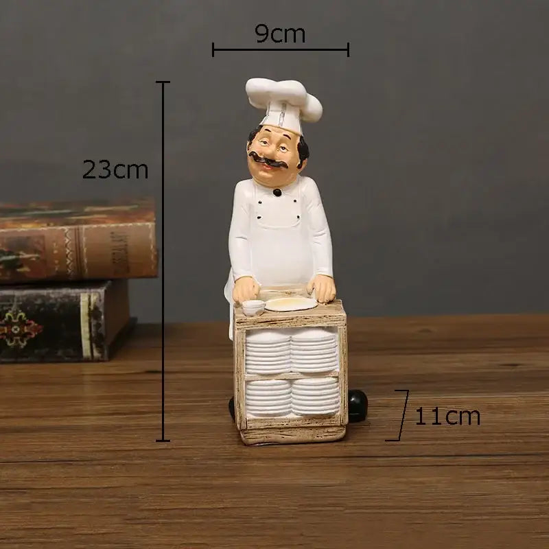 A charming white bearded chef resin figure with tall toque standing behind a rustic wooden cart stacked with plates