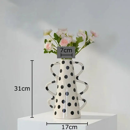 A tall white ceramic vase with black polka dot design and sculpted handles holding pink and cream flowers