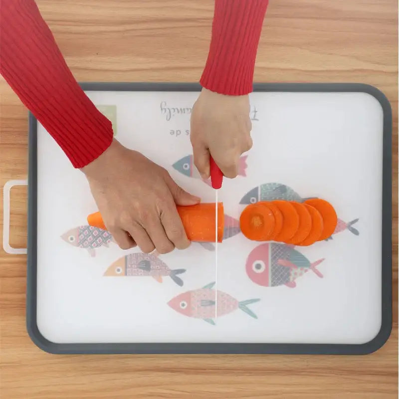 White and gray fish design cutting board with colorful pattern and red-handled knife slicing carrot rounds on chopping block decorbites dual-use cutting