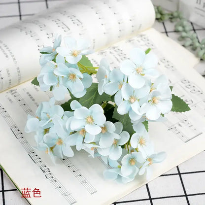 A delicate light blue hydrangea silk flower bouquet with layered petals and yellow centers in a white begonia silk flower vase