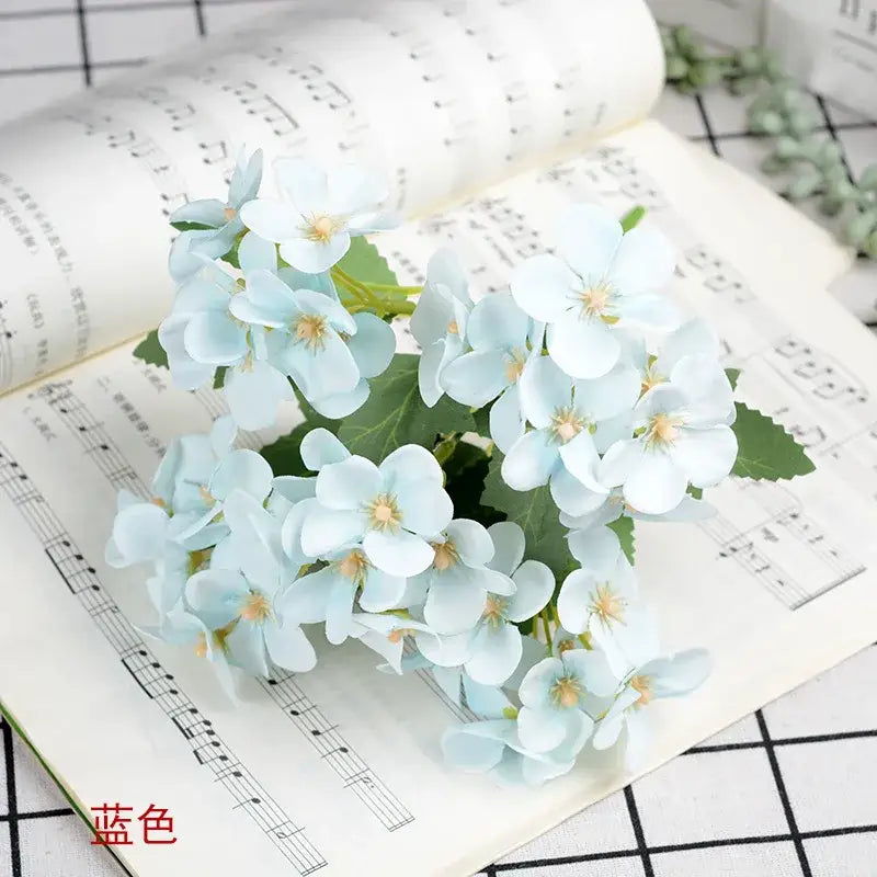 A delicate light blue hydrangea silk flower bouquet with layered petals and yellow centers in a white begonia silk flower vase