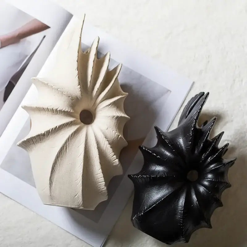 Two conch shape ceramic vases in matte off-white and glossy black for artificial flower arrangements on magazine