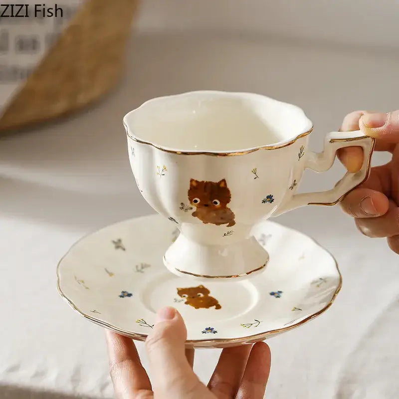 White ceramic teacup and saucer with gold trim featuring a cartoon kitten pattern surrounded by blue flowers