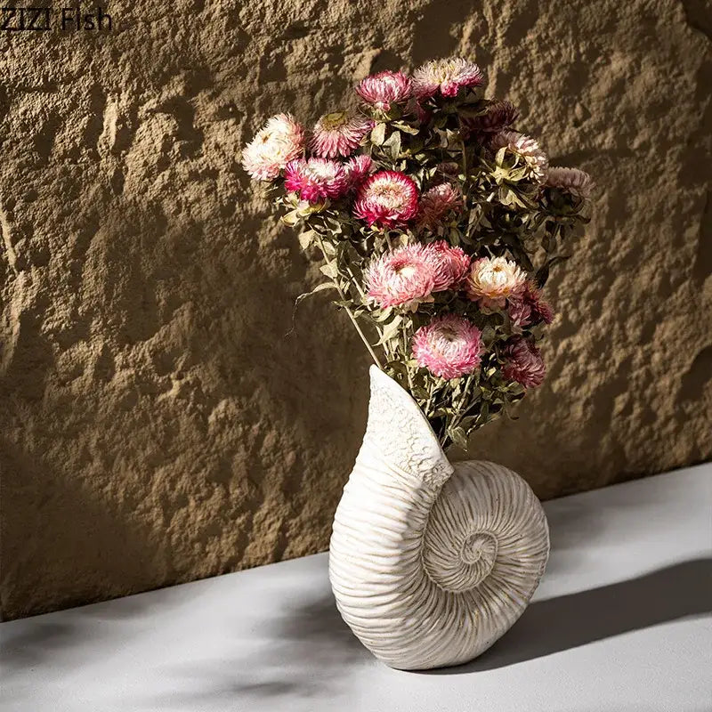 White nautilus conch ceramic vase by decorbites with spiral seashell shape holding dried pink and cream flowers for decor