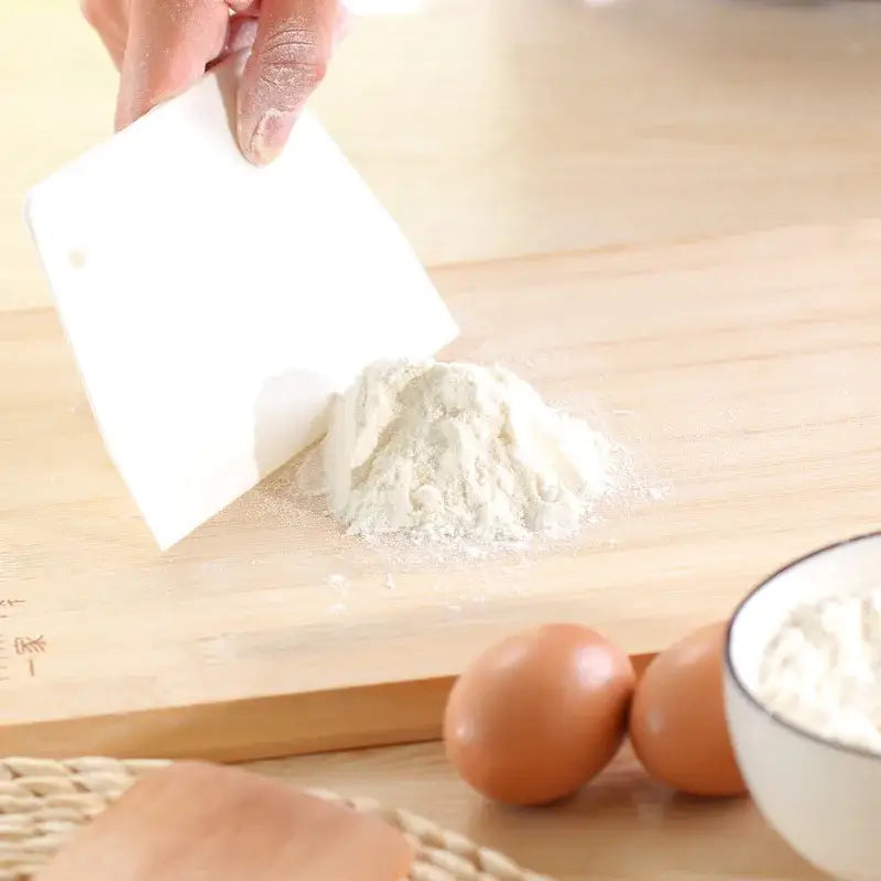 White plastic dough cream cake cutter spatula gathering flour on wooden cutting board for kitchen baking tasks