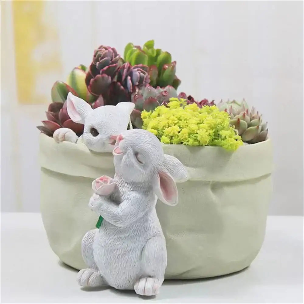 A white resin bunny succulent planter pot holding a pink rose next to a light green decorbites bunny succulent garden