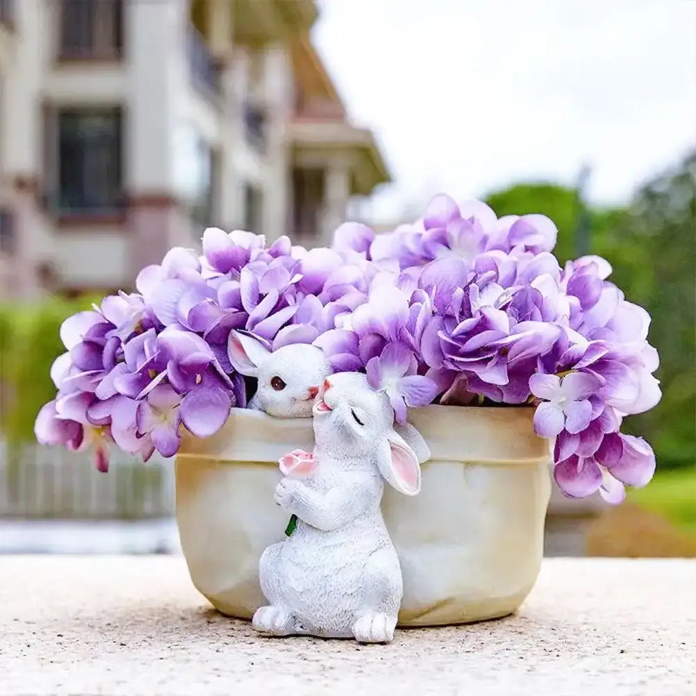 A white resin decorbites bunny succulent planter pot with pink accents beside a cream ceramic pot of purple hydrangeas