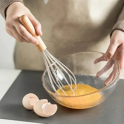 Wooden-handled metal whisk beating eggs in glass bowl from white silicone kitchen cookware set with wooden handles