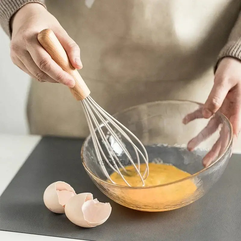 Wooden-handled metal whisk beating eggs in glass bowl from white silicone kitchen cookware set with wooden handles