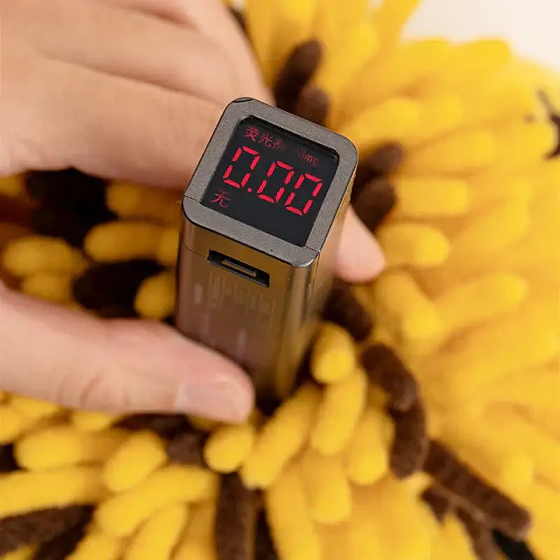 A compact metallic black digital device with red led display and usb port next to a yellow and black bee shape chenille hand towel