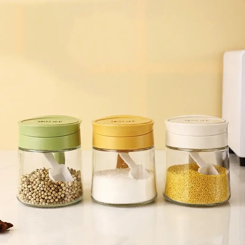 Set of three clear glass seasoning jars with green yellow and white lids each containing a plastic scoop spoon
