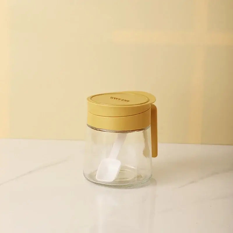 Yellow flip top glass seasoning jar with mustard yellow lid, handle, and integrated white spoon for kitchen use