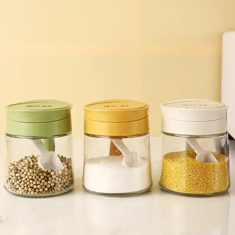Three clear glass seasoning jars with green yellow and white lids each containing different seasonings and a yellow flip top glass seasoning jar with spoon